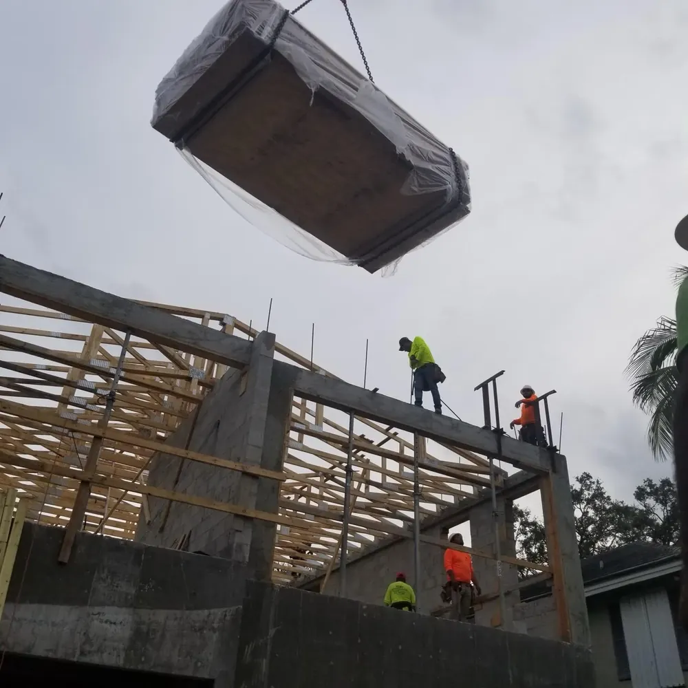 A crane lifting a wrapped pallet of roofing materials to workers on the second story of a commercial construction site managed by Booster Construction in Miami.